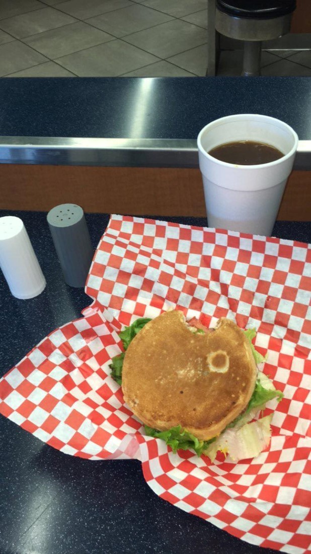 Photo of a greasy American burger sitting on top of red and white checkered paper. Next to the burger is a coffee in a styrofoam cup.