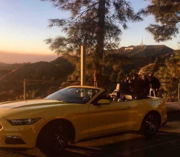 A luxurious yellow convertible parked by a look-out. In the background is the Hollywood sign.